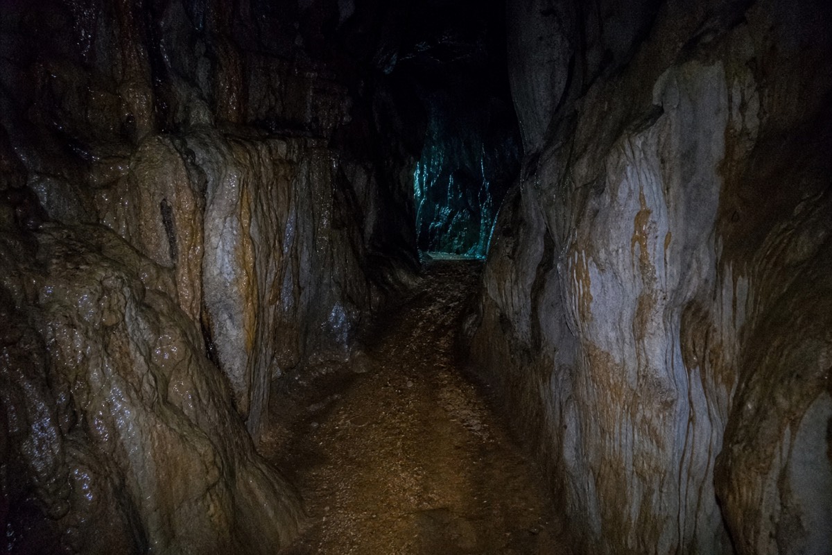 Looking down a dark limestone passage towards a faint blue-green glow around the corner with wet rock walls close on both sides