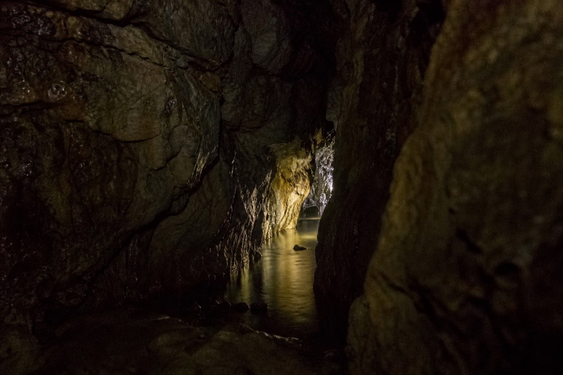 Another view at Waitomo: a dark limestone cave passage narrowing towards a lit doorway-sized gap ahead with deep shadow either side