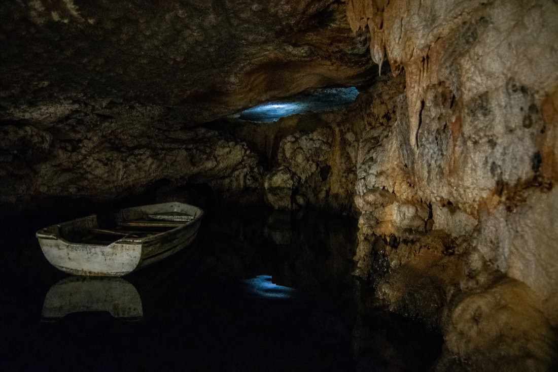 Another view at Waitomo: a small quiet cave chamber with a low ceiling and one empty inflatable boat on still black water beneath faint blue-green light