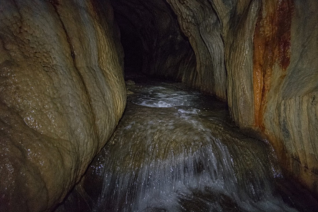 Another view at Waitomo: a dark limestone cave passage with fast shallow water running over a rock lip under weak side lighting
