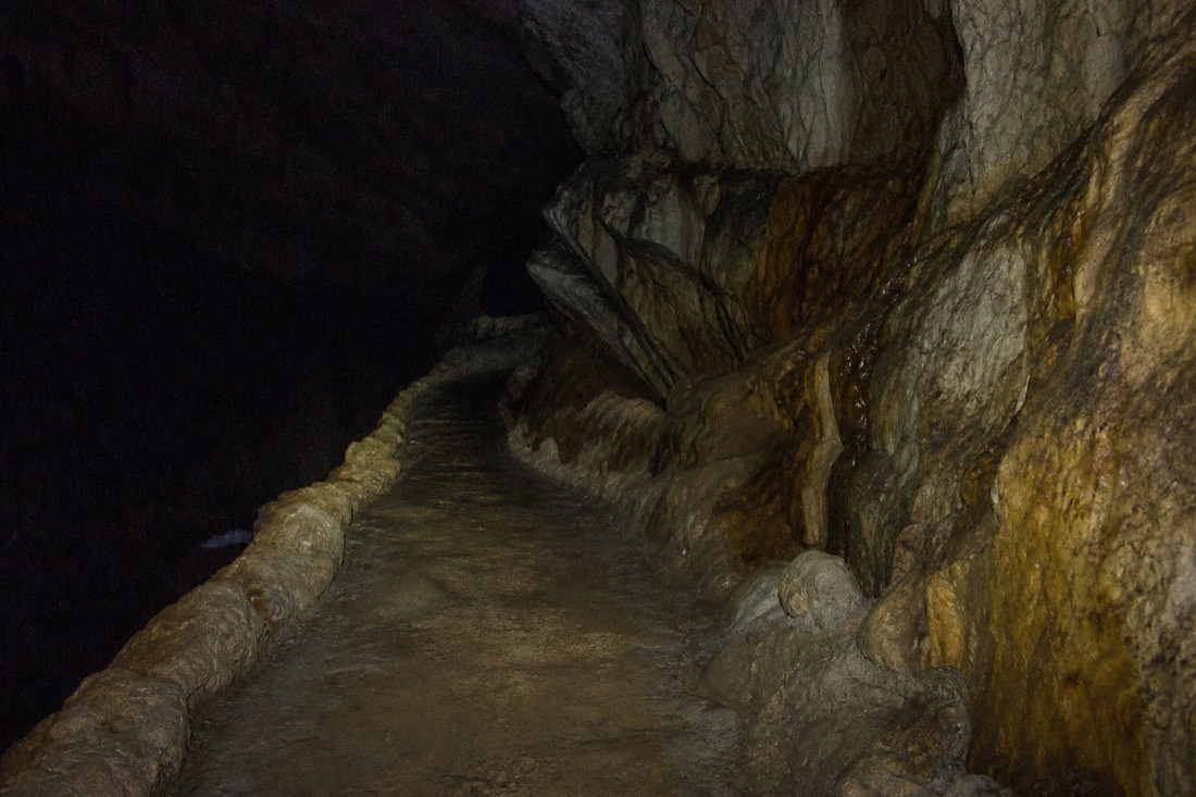 Another view at Waitomo: looking down a dark limestone passage towards a faint blue-green glow around the corner with wet rock walls close on both sides