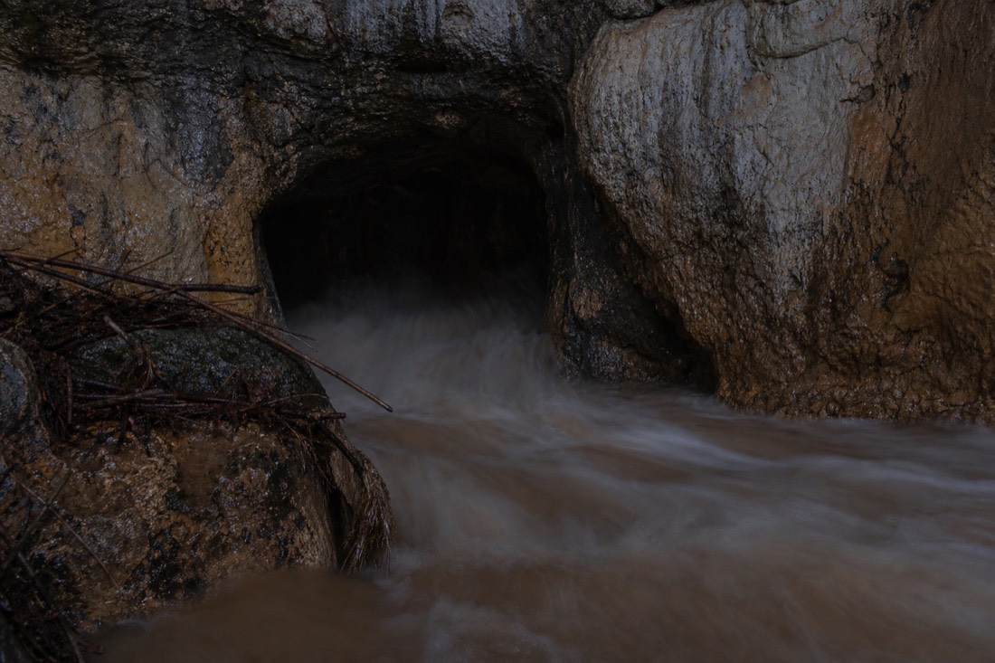Another view at Waitomo: a brown swollen river squeezing into a limestone cave mouth after heavy rain with debris caught on a rock lip