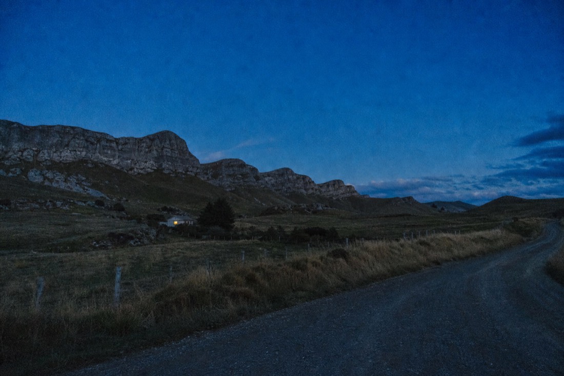 Another view at Waitomo: a small New Zealand rural valley at dusk with scattered farm buildings and limestone bluffs behind under a deep blue sky